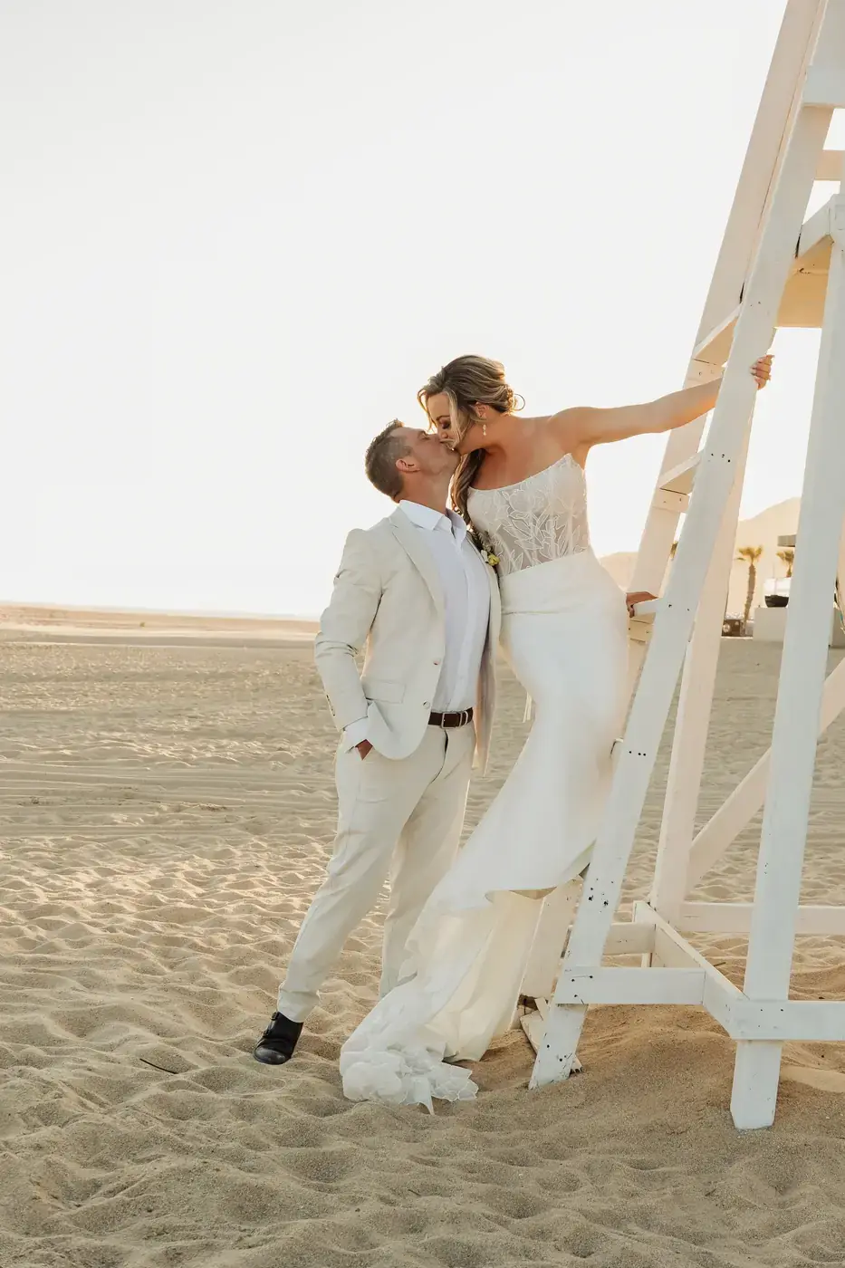 Bride and groom kissing against a white lifeguard tower on the beach at golden hour in Cabo