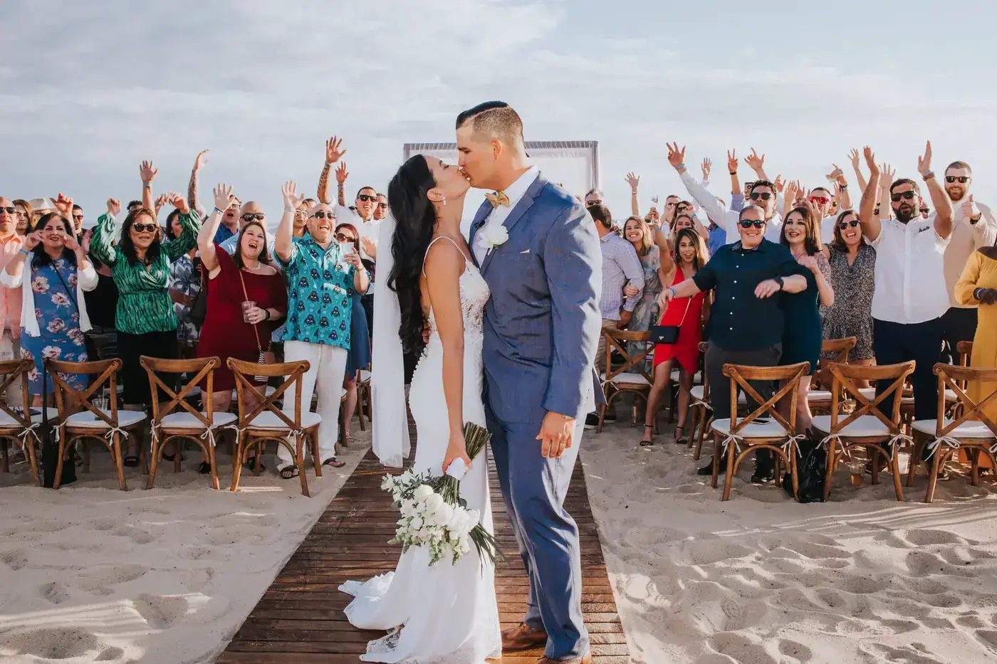 Couple at the ceremony arch while guests raise their hands in celebration in Cabo