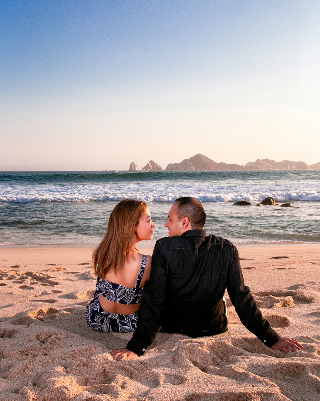 Couple sitting on the sand at Playa Monumentos in Cabo San Lucas with El Arco on the horizon at golden hour